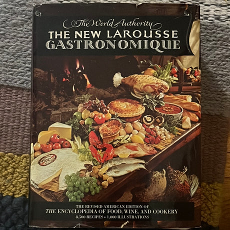 The World Authority The New Larousse Gastronomique book on a rug, showcasing its cover featuring culinary elements and food imagery, emphasizing its encyclopedic nature.