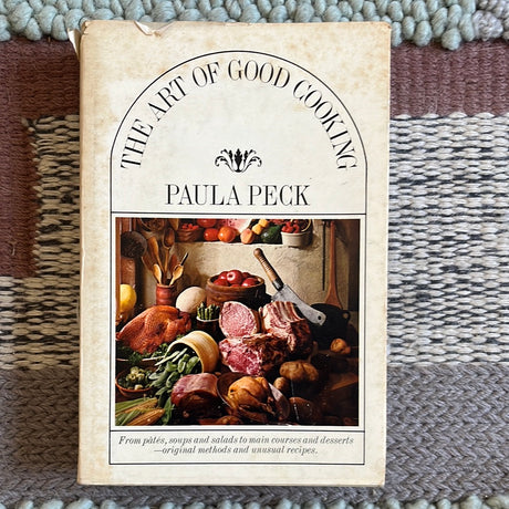 Cover of The Art of Good Cooking cookbook on a rug, featuring recipes by Paula Peck with a knife and meat nearby.