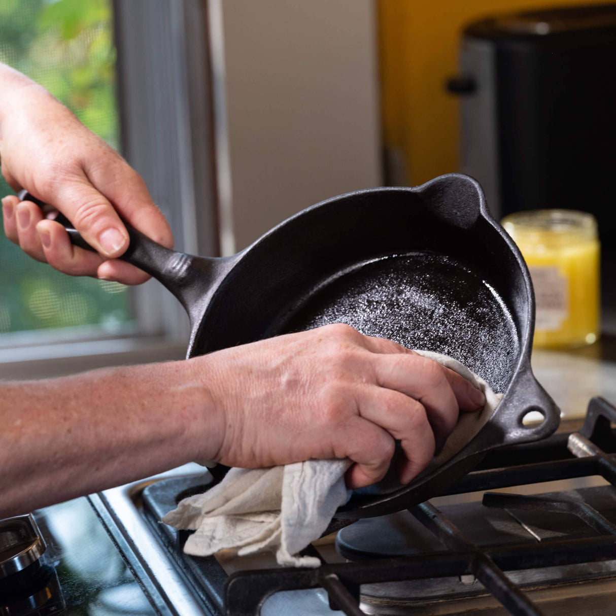 Person seasoning a small cast-iron skillet with cloth using Cast Iron Seasoning Oil over a gas stovetop, enhancing cookware longevity and non-stick surface.