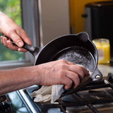 Person seasoning a small cast-iron skillet with cloth using Cast Iron Seasoning Oil over a gas stovetop, enhancing cookware longevity and non-stick surface.
