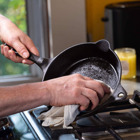 Person seasoning a small cast-iron skillet with cloth using Cast Iron Seasoning Oil over a gas stovetop, enhancing cookware longevity and non-stick surface.