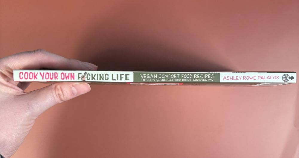 Cookbook spine reading Cook Your Own F*cking Life by Ashley Rowe Palafox, held in hand, promoting vegan comfort food recipes and community building.