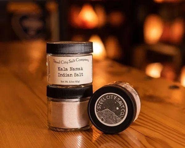 Three Steel City Salt Company jars of Kala Namak Indian Salt on wood; two stacked, one lying down, highlighting its vegan-friendly, egg-flavored culinary use.