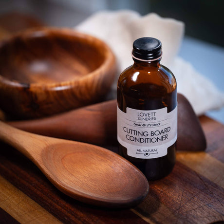 Coconut Oil Cutting Board Conditioner in amber glass bottle on a wooden board, with a wooden spoon in front, and a bowl and towel behind.