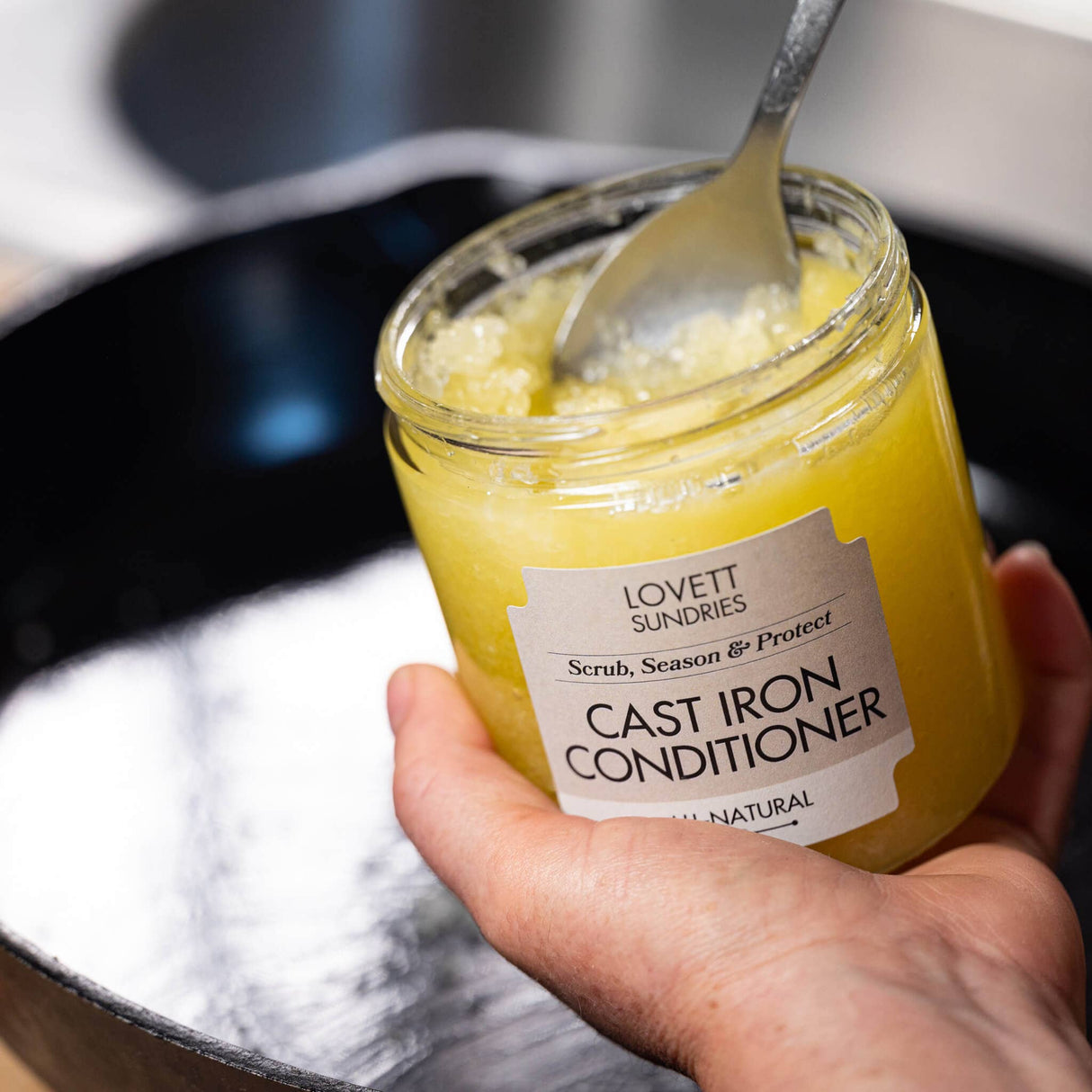 Person holding Lovett Sundries Cast Iron Conditioner Scrub and Seasoning jar with spoon over cast-iron pan, highlighting its use for cleaning and protecting cookware.